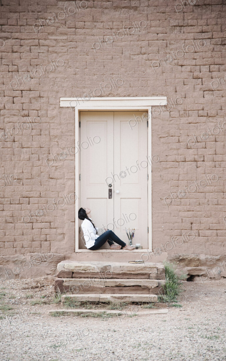 Young woman with long brown hair sitting on the ground outside the front door of a…