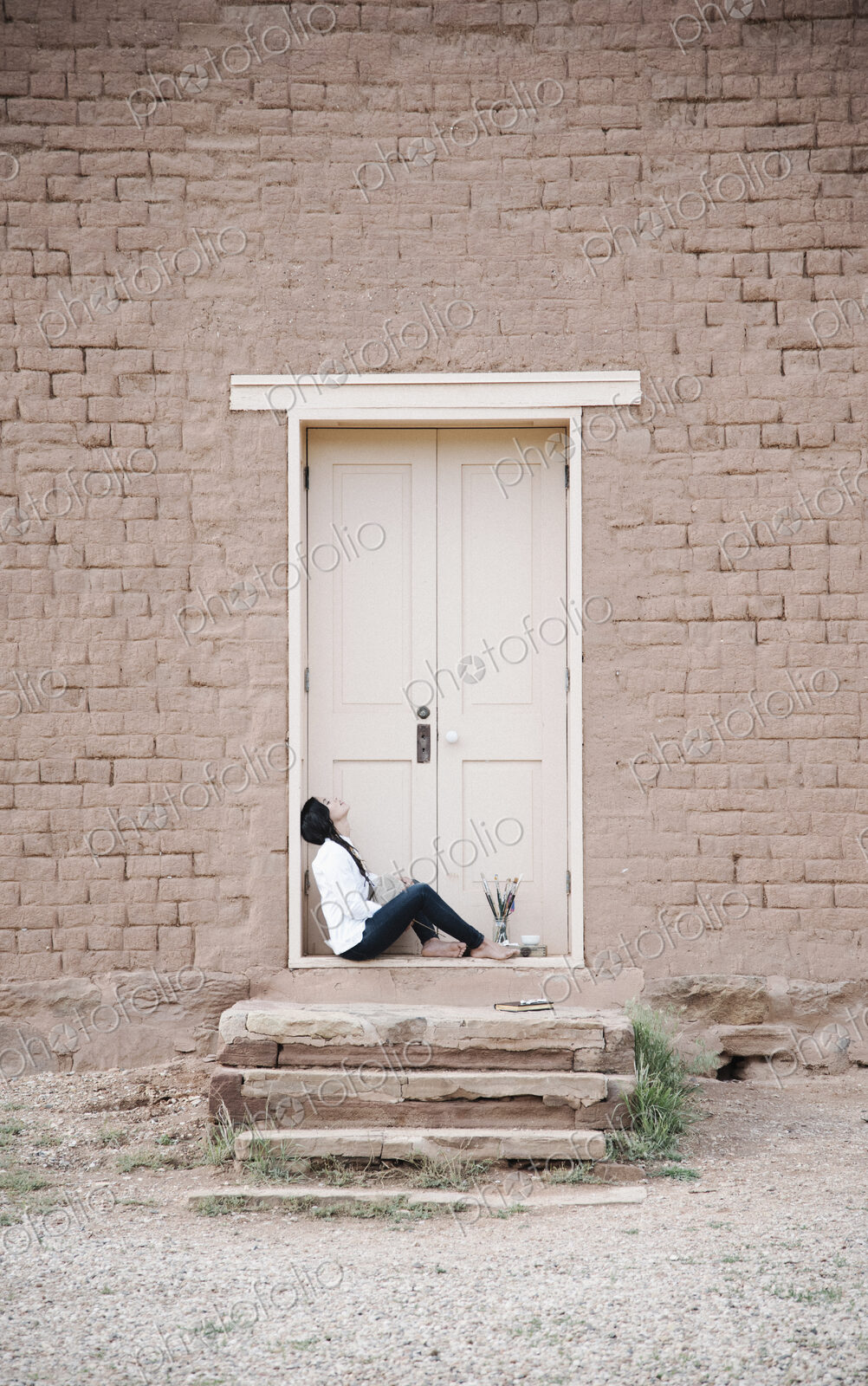 Young woman with long brown hair sitting on the ground outside the front door of a…