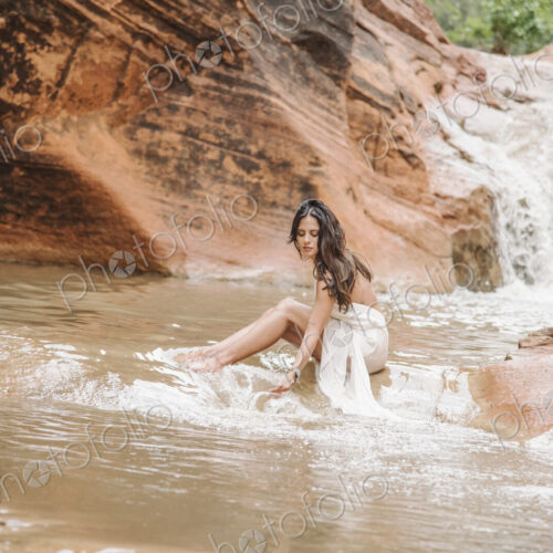 Young woman with long brown hair, wearing a long white dress, sitting on a rock in a… Young woman with long brown hair, wearing a long white dress, sitting on a rock in a…