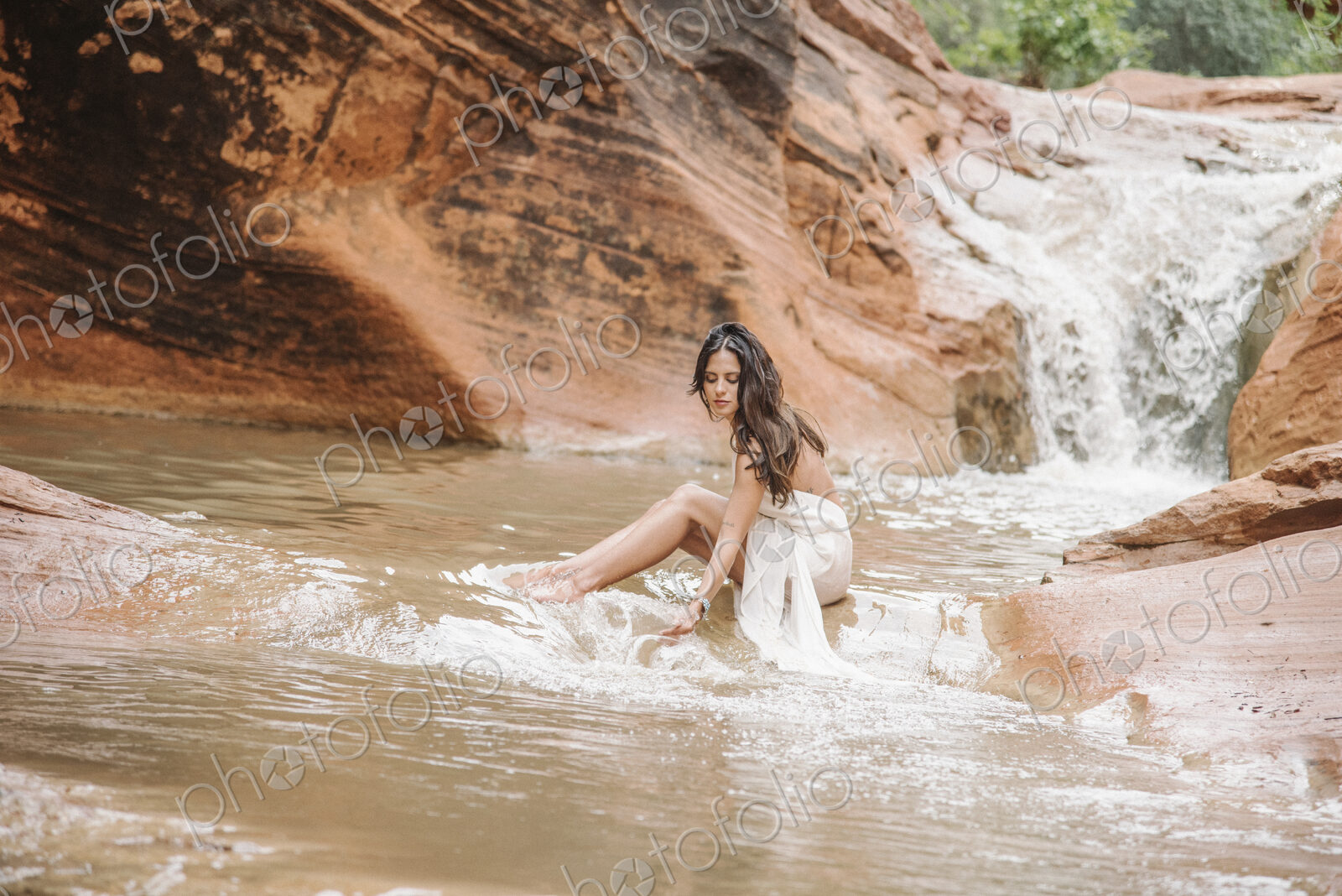 Young woman with long brown hair, wearing a long white dress, sitting on a rock in a…