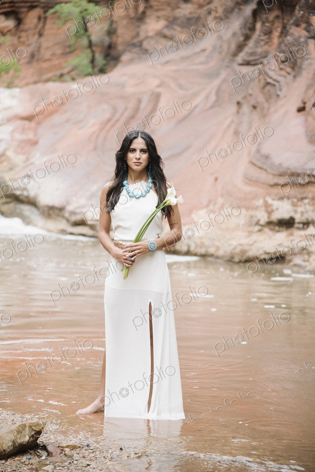 Young woman with long brown hair, wearing a long white dress, standing by a river,…