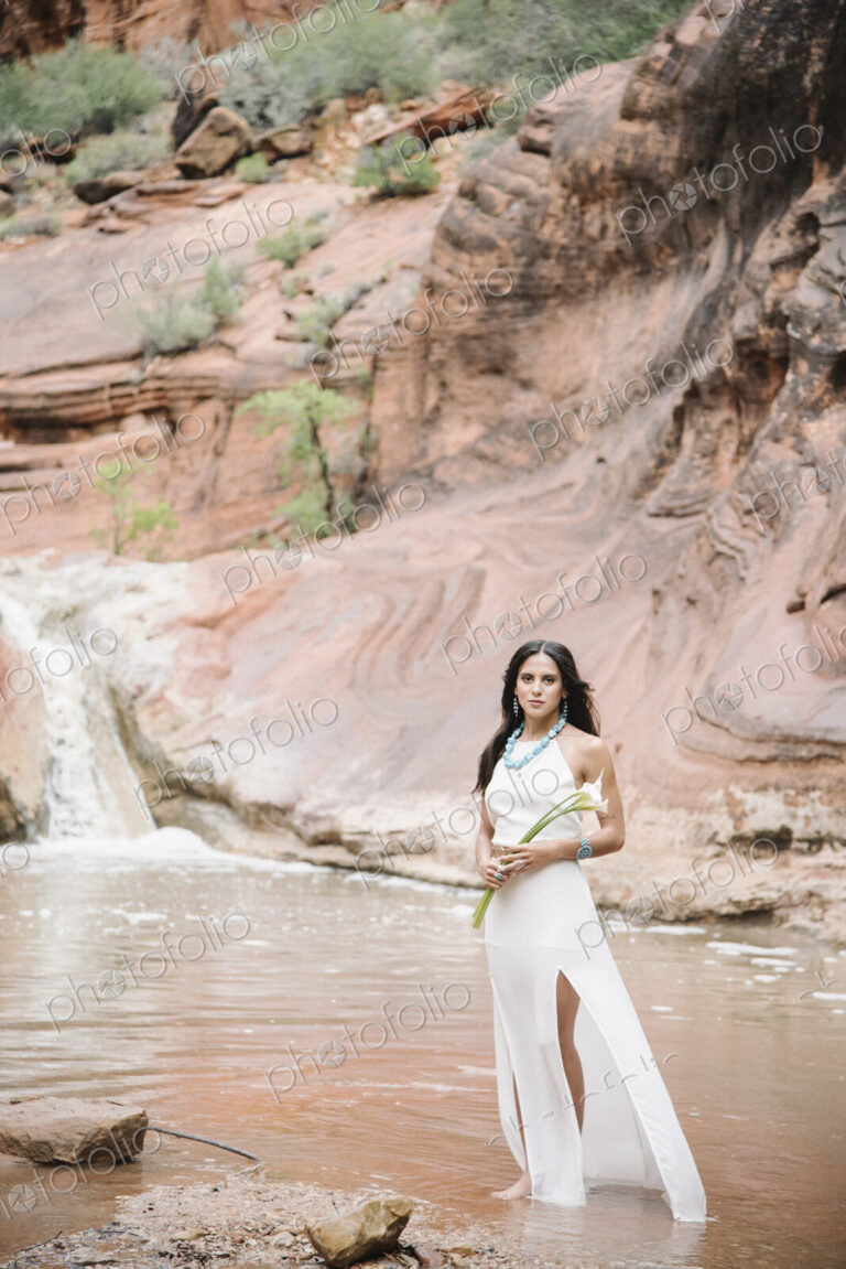 Young woman with long brown hair, wearing a long white dress, standing by a river,…
