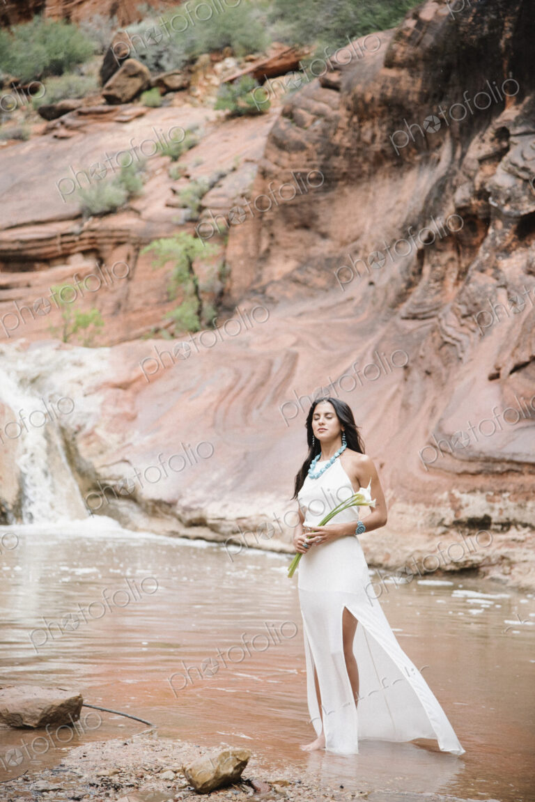 Young woman with long brown hair, wearing a long white dress, standing by a river,…