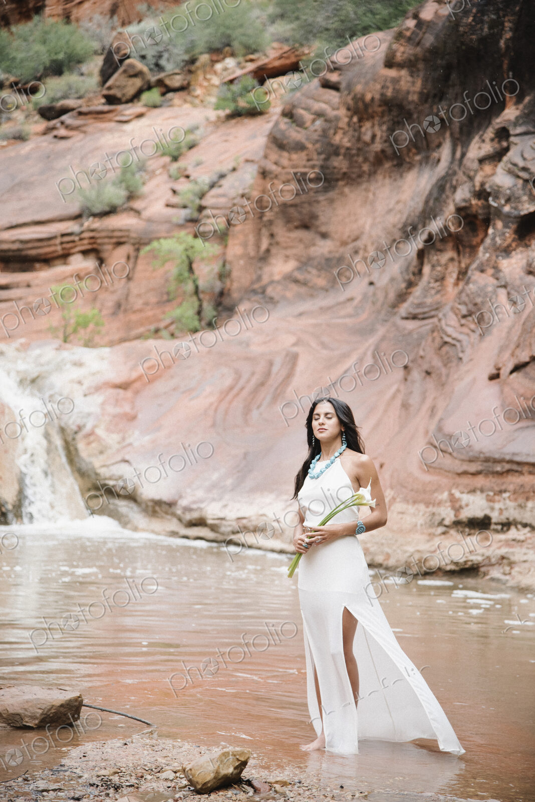 Young woman with long brown hair, wearing a long white dress, standing by a river,…