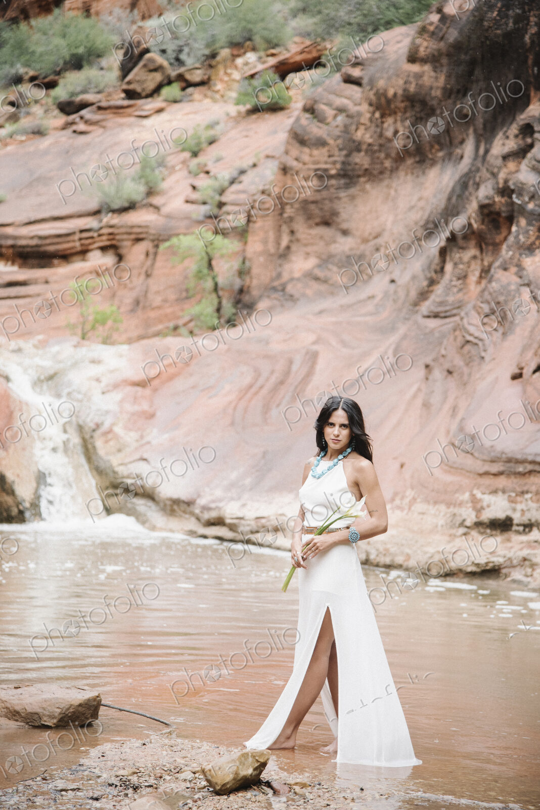Young woman with long brown hair, wearing a long white dress, standing by a river,…