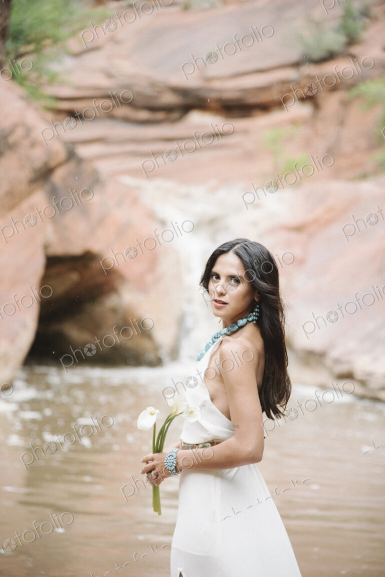 Young woman with long brown hair, wearing a long white dress, standing by a river,…