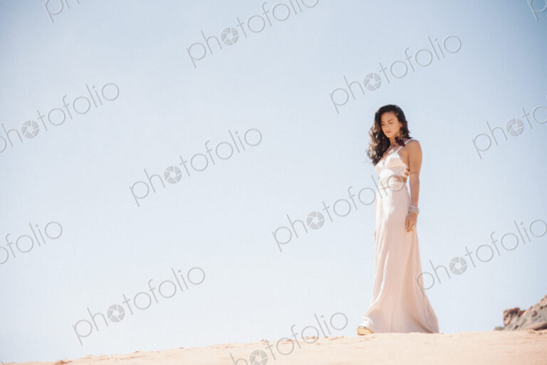 Young woman with long brown hair, wearing a long white dress, standing in a desert.