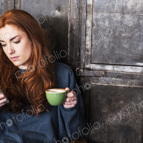 Young woman with long red hair sitting at table, holding cup of coffee, checking her…