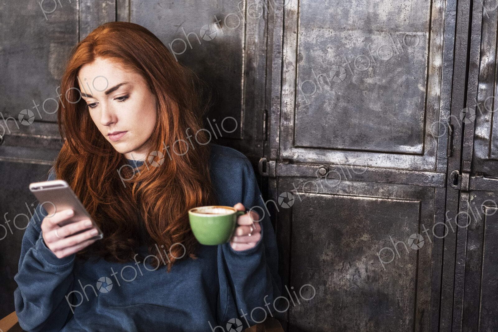 Young woman with long red hair sitting at table, holding cup of coffee, checking her…