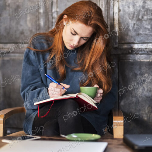 Young woman with long red hair sitting at table, holding notebook and cup of coffee. Young woman with long red hair sitting at table, holding notebook and cup of coffee.