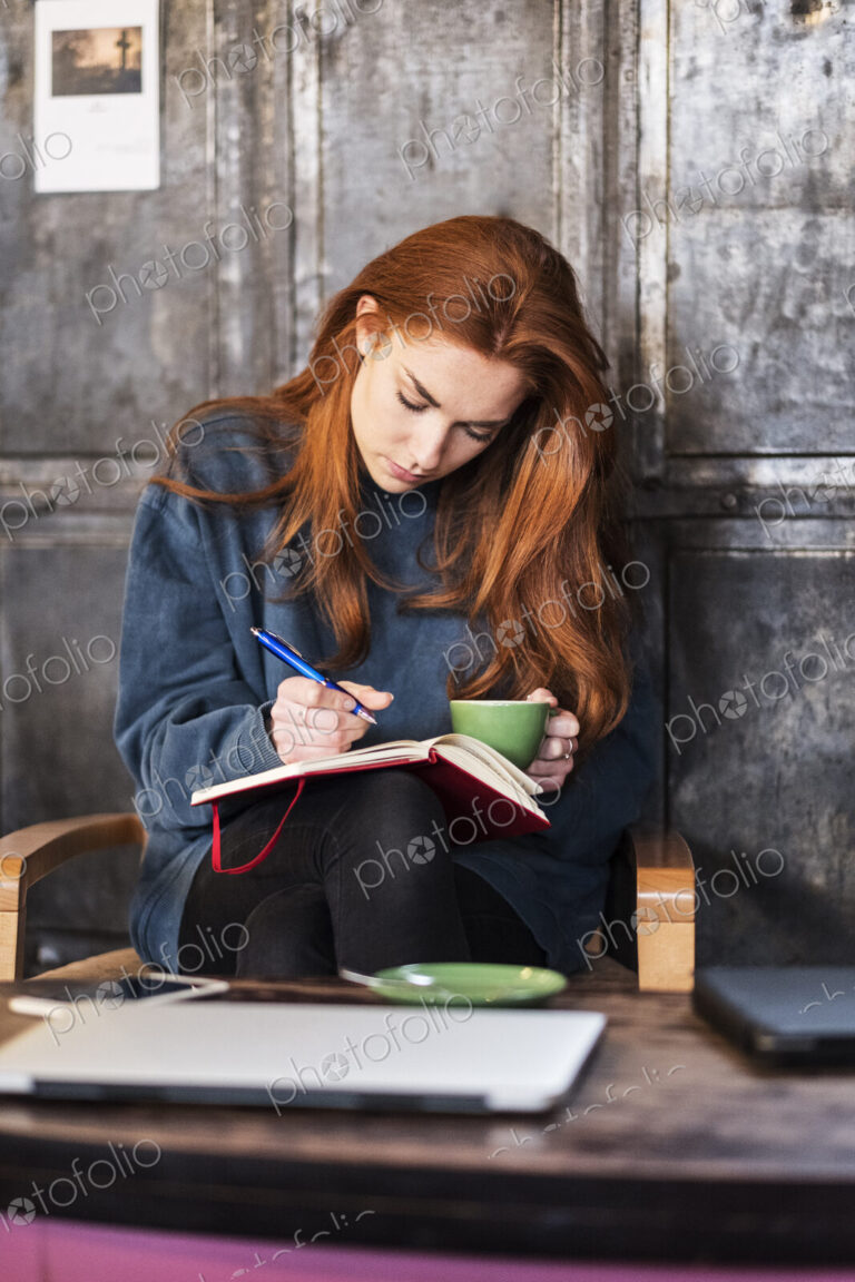 Young woman with long red hair sitting at table, holding notebook and cup of coffee.