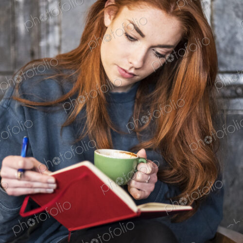 Young woman with long red hair sitting at table, holding notebook and cup of coffee. Young woman with long red hair sitting at table, holding notebook and cup of coffee.