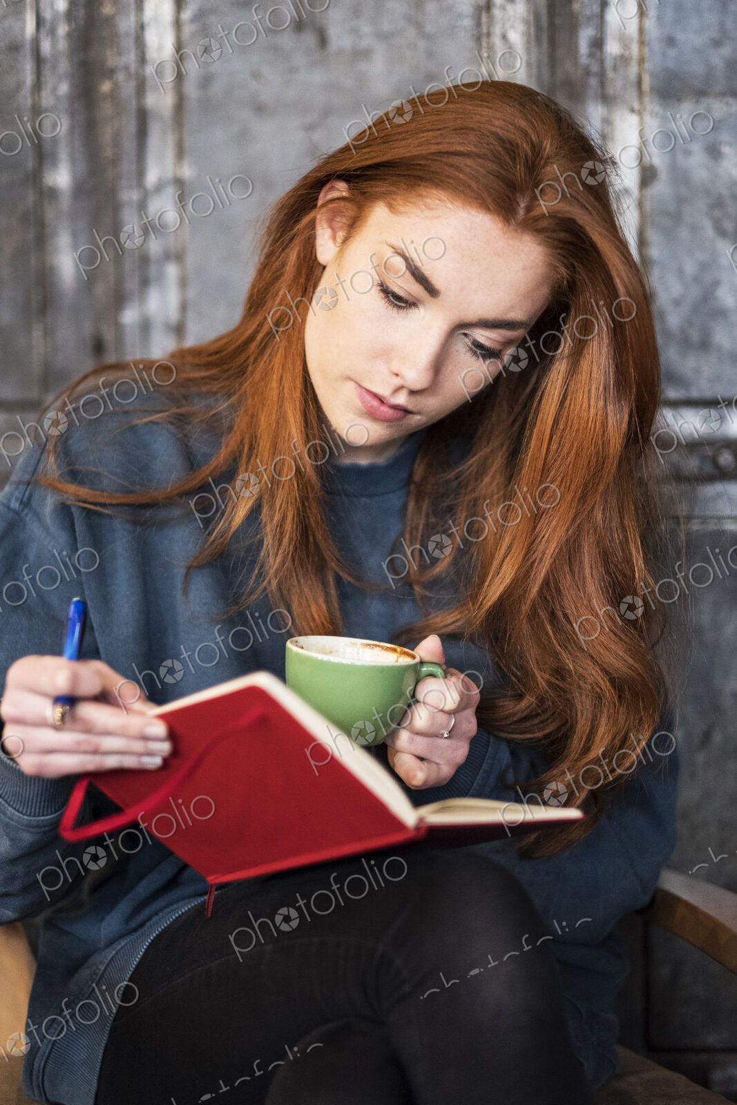 Young woman with long red hair sitting at table, holding notebook and cup of coffee.