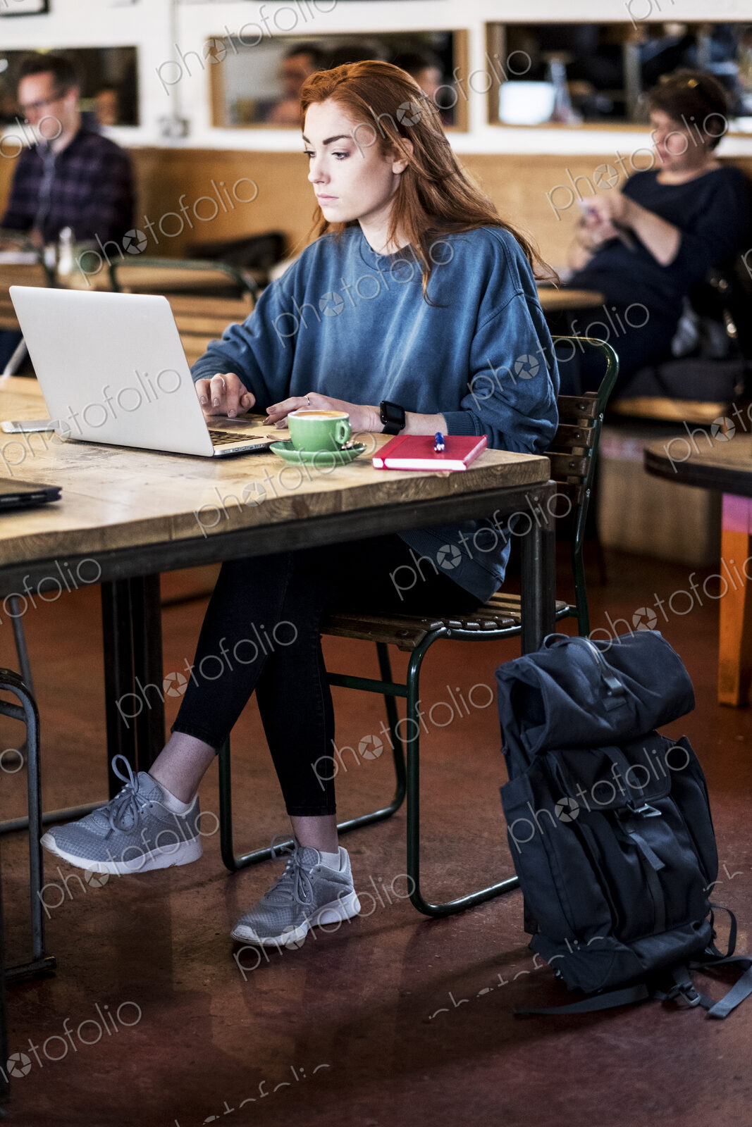 Young woman with long red hair sitting at table, working on laptop computer.