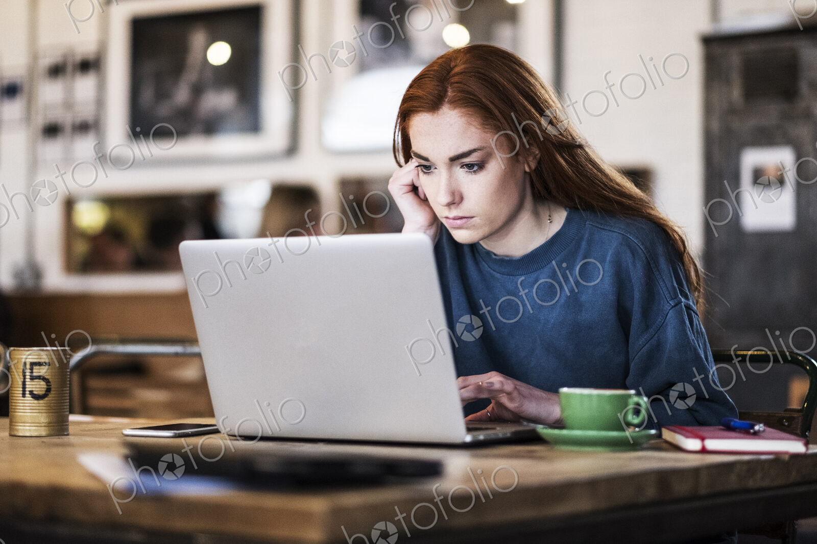 Young woman with long red hair sitting at table, working on laptop computer.