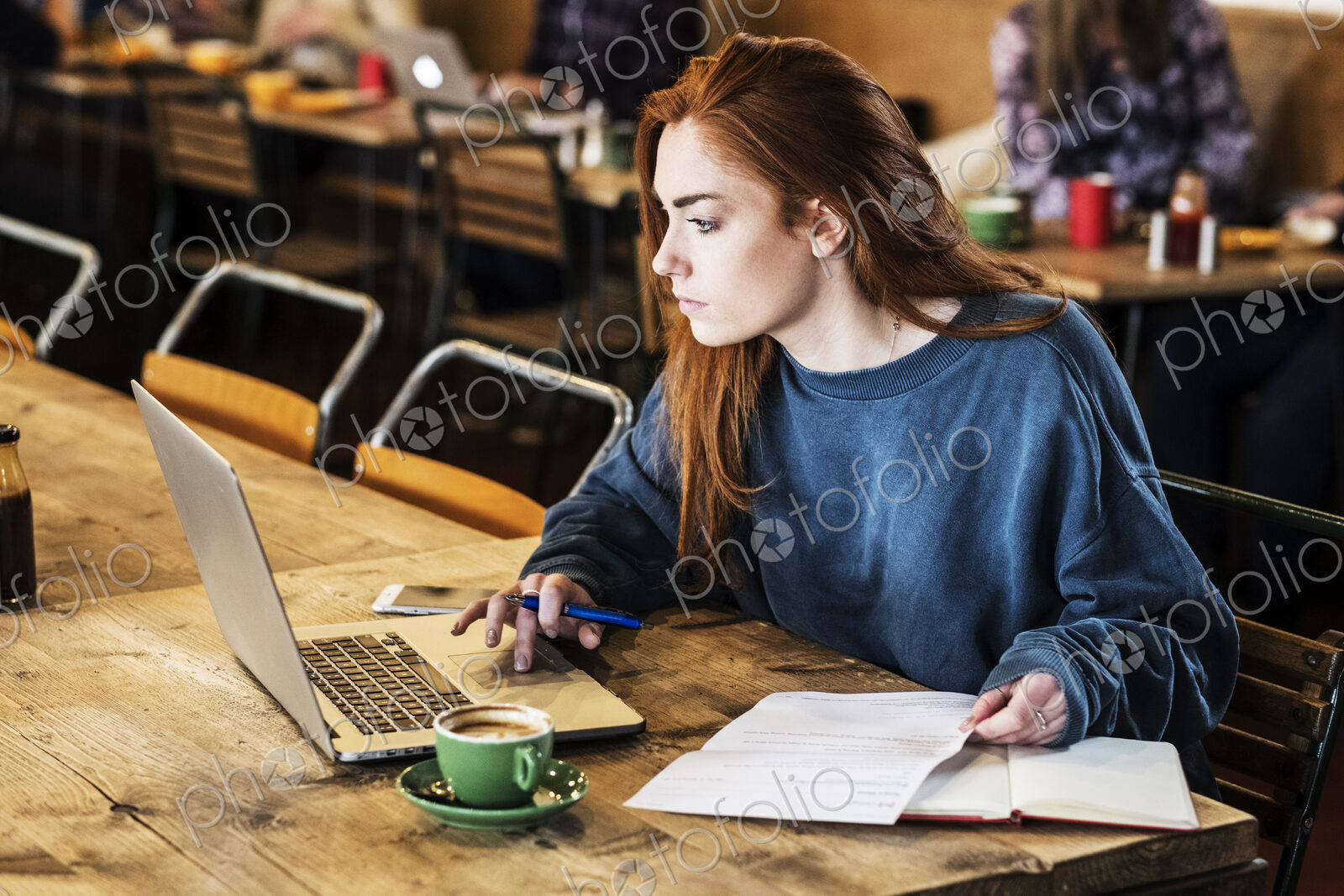 Young woman with long red hair sitting at table, working on laptop computer.