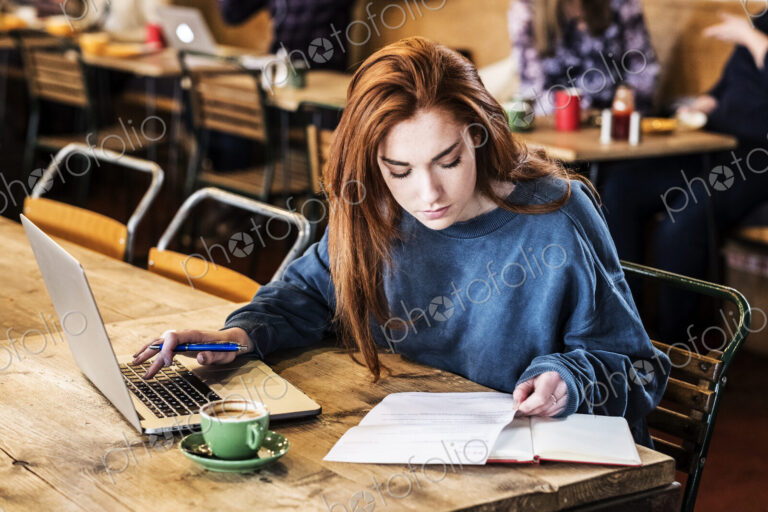 Young woman with long red hair sitting at table, working on laptop computer.