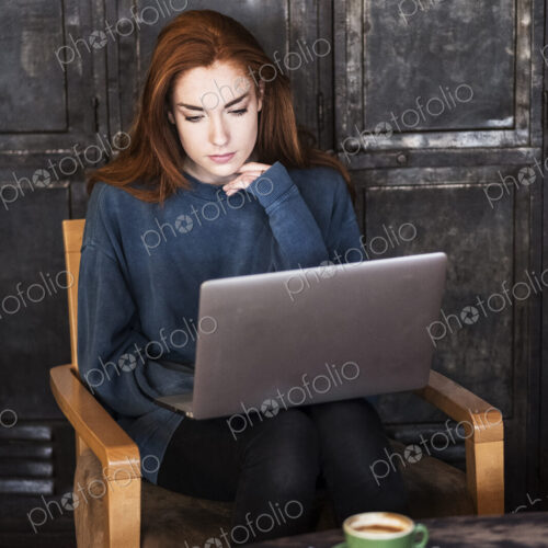 Young woman with long red hair sitting at table, working on laptop computer.
