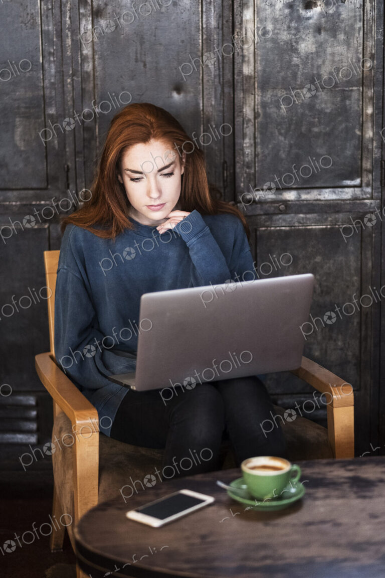Young woman with long red hair sitting at table, working on laptop computer.