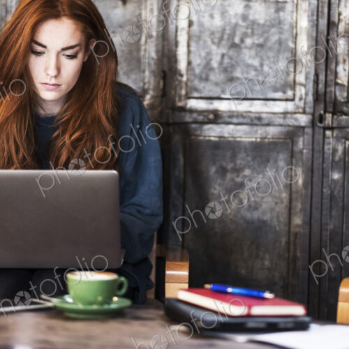 Young woman with long red hair sitting at table, working on laptop computer.