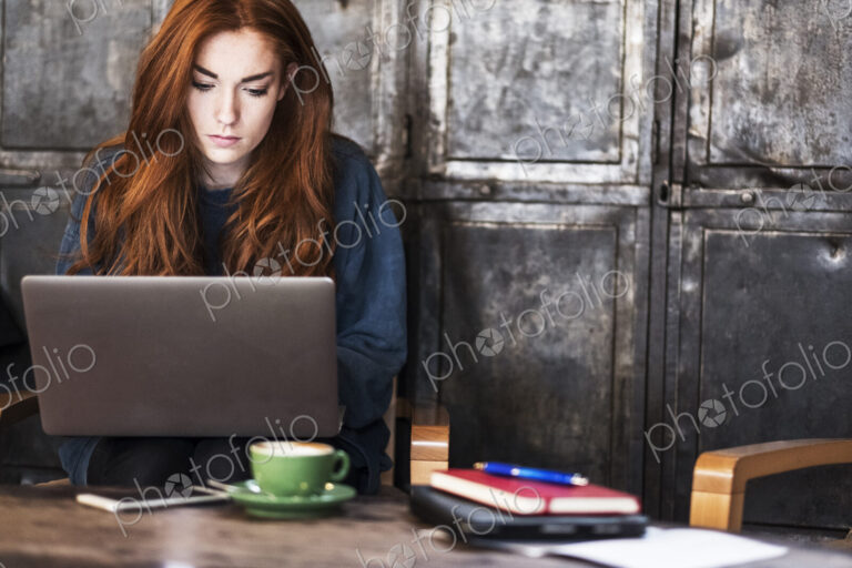 Young woman with long red hair sitting at table, working on laptop computer.