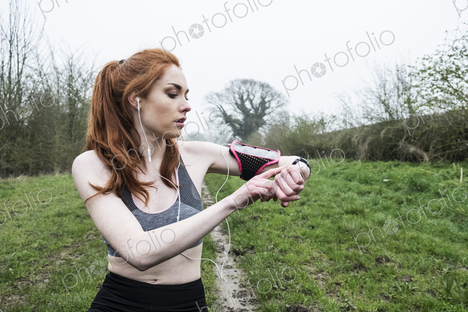 Young woman with long red hair wearing sportswear, exercising outdoors, checking her…