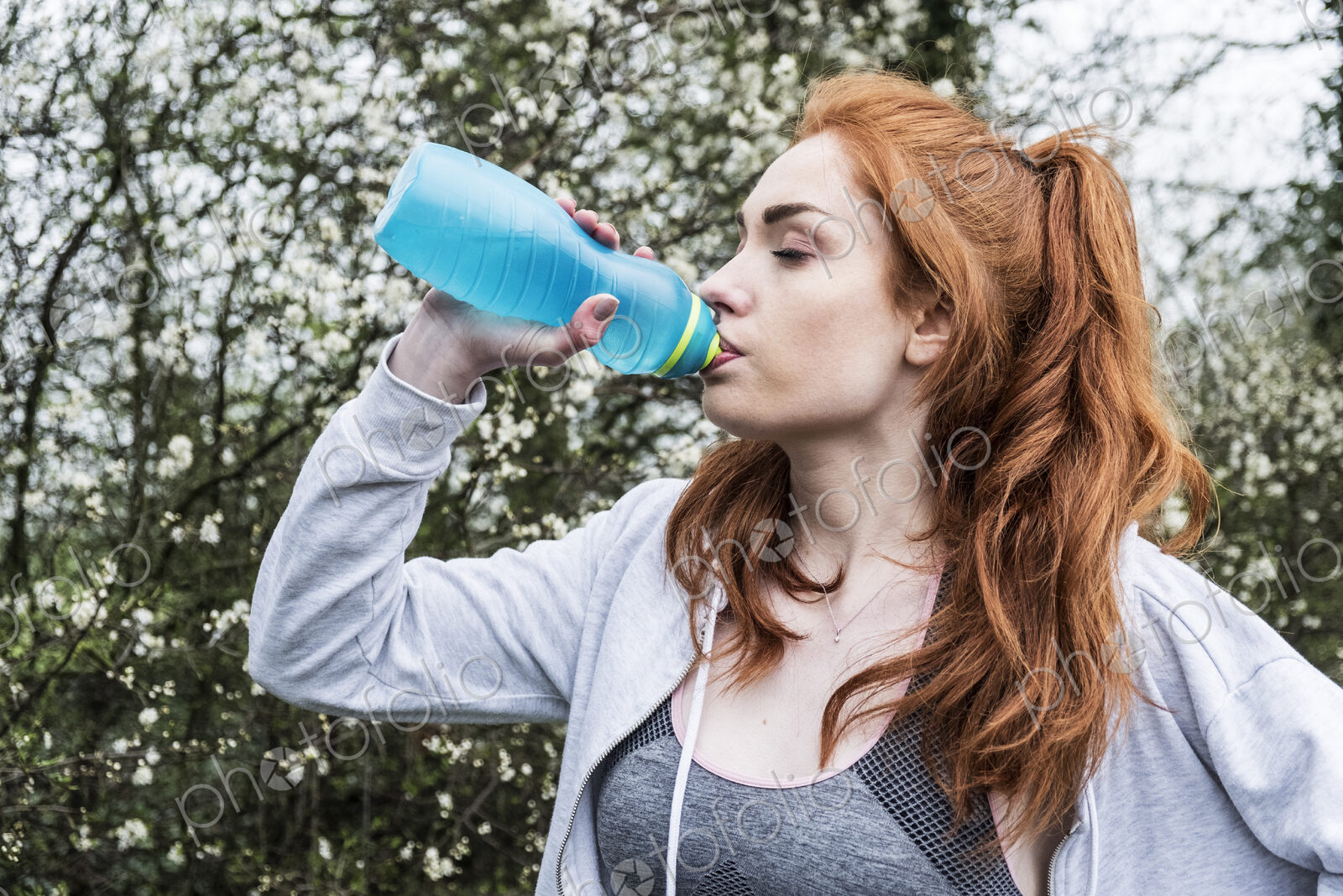 Young woman with long red hair wearing sportswear, exercising outdoors, drinking from…