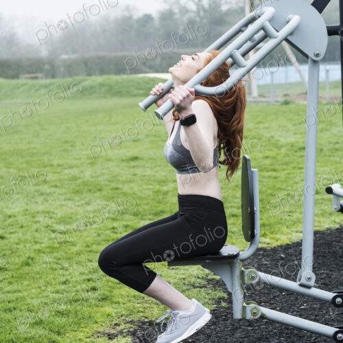 Young woman with long red hair wearing sportswear, using outdoor exercise machine. Young woman with long red hair wearing sportswear, using outdoor exercise machine.