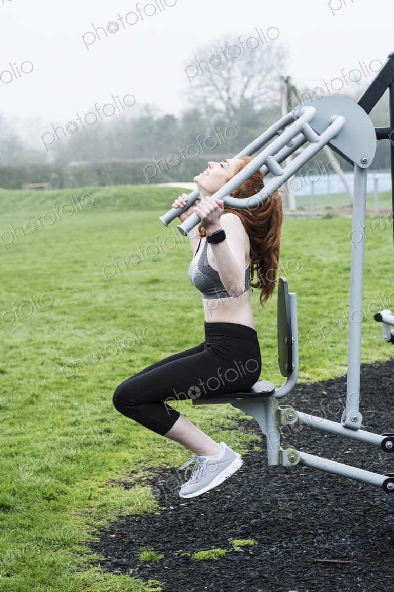 Young woman with long red hair wearing sportswear, using outdoor exercise machine.
