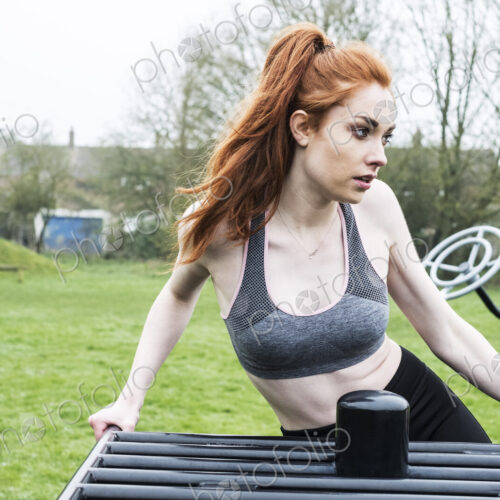 Young woman with long red hair wearing sportswear, using outdoor exercise machine.