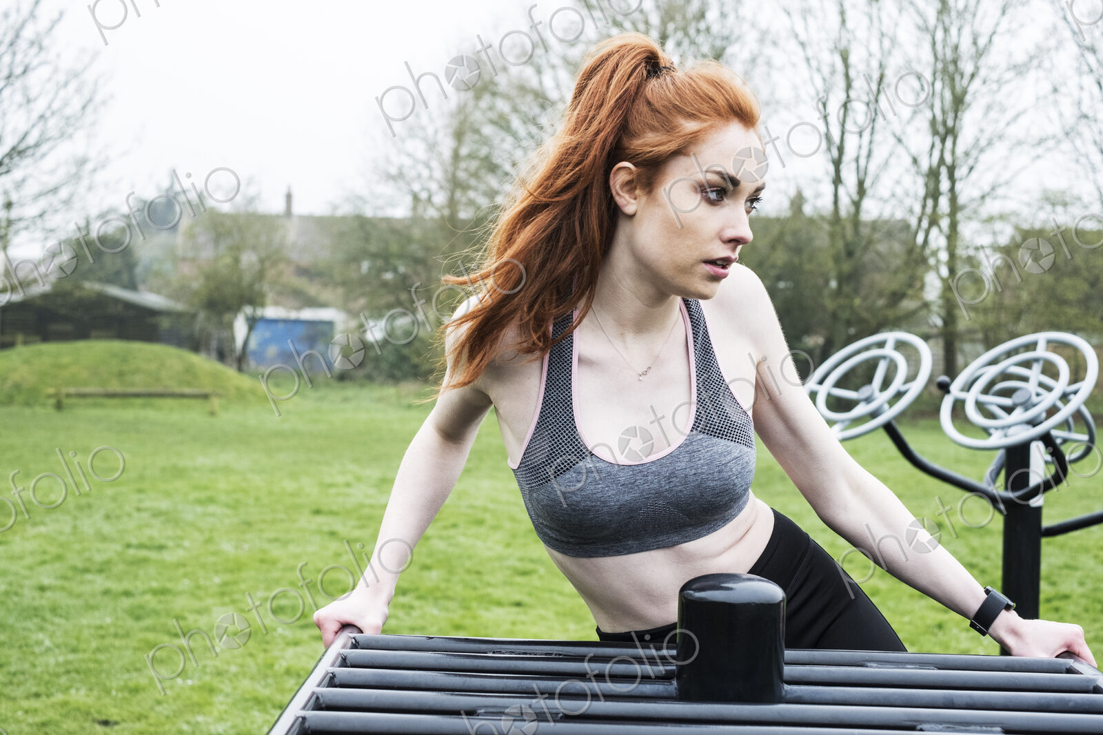 Young woman with long red hair wearing sportswear, using outdoor exercise machine.