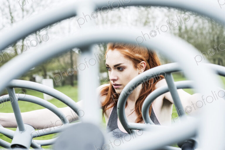 Young woman with long red hair wearing sportswear, using outdoor exercise machine.