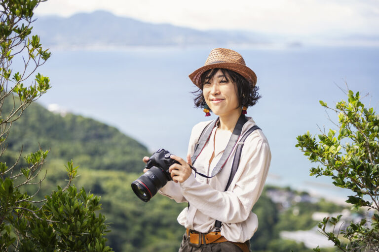 Japanese woman wearing hat and carrying backpack and camera standing on a cliff, ocean…