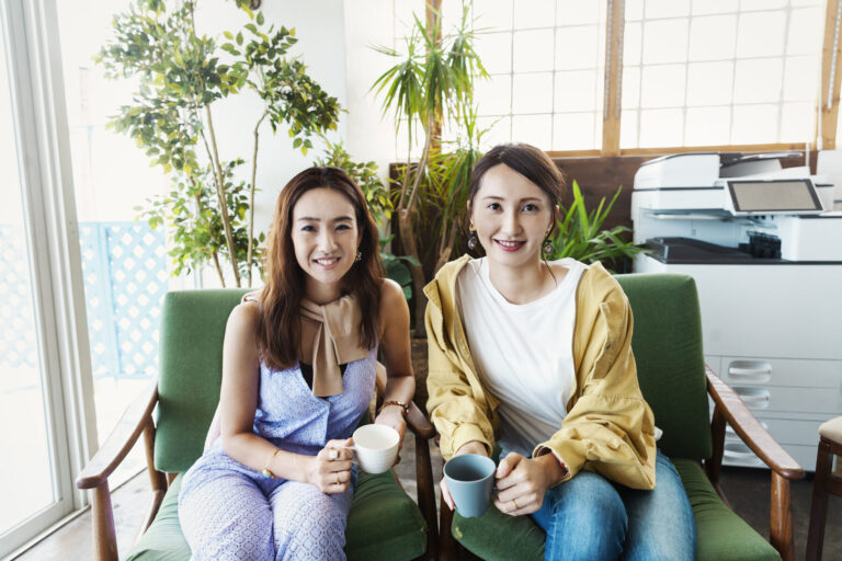 Two female Japanese professionals sitting in a co-working space, smiling at camera.