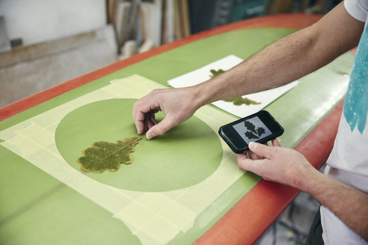 People category — Man creating a design on surfboard laying an oak leaf on the board, using mobile phone… Man creating a design on surfboard laying an oak leaf on the board, using mobile phone…